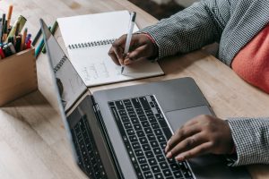 Crop unrecognizable African American female in formal wear writing notes in notepad while working on laptop — Explore the Inc