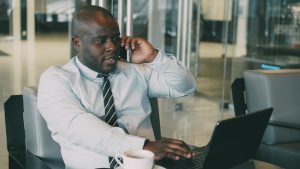 Professional man multitasking, talking on the phone while using a laptop in an office. — IronFX Reviews: A Reliable Broker Co